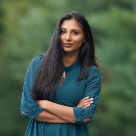 Pooja Agrawal in a blue dress with trees in the background with her arms crossed looking attentively at the camera